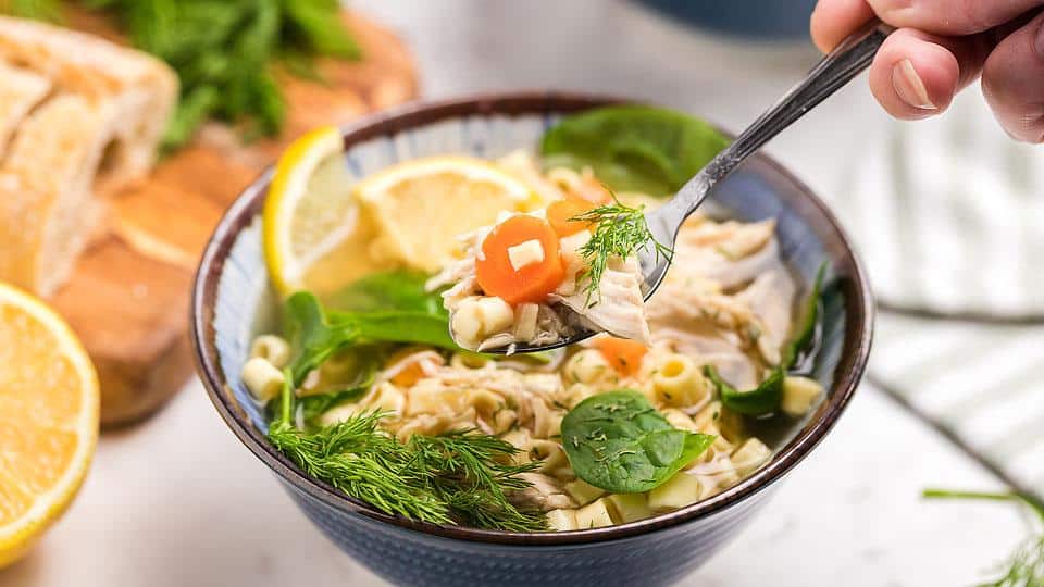 A bowl of chicken noodle soup with carrots, spinach, dill, and lemon slices, with a spoon lifting soup above the bowl. Bread and lemon are in the background.