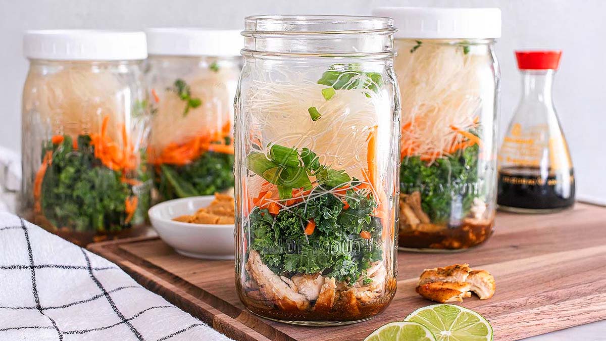 Four mason jars filled with layers of noodles, shredded carrots, greens, and chicken sit on a wooden board, with a soy sauce bottle and sliced lime nearby.