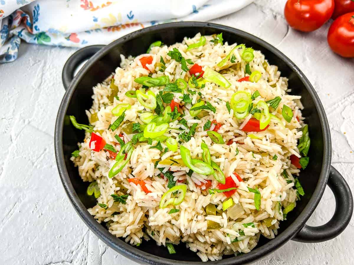 A black bowl filled with cooked rice mixed with chopped green onions, red bell peppers, and herbs, placed on a white surface with tomatoes in the background.