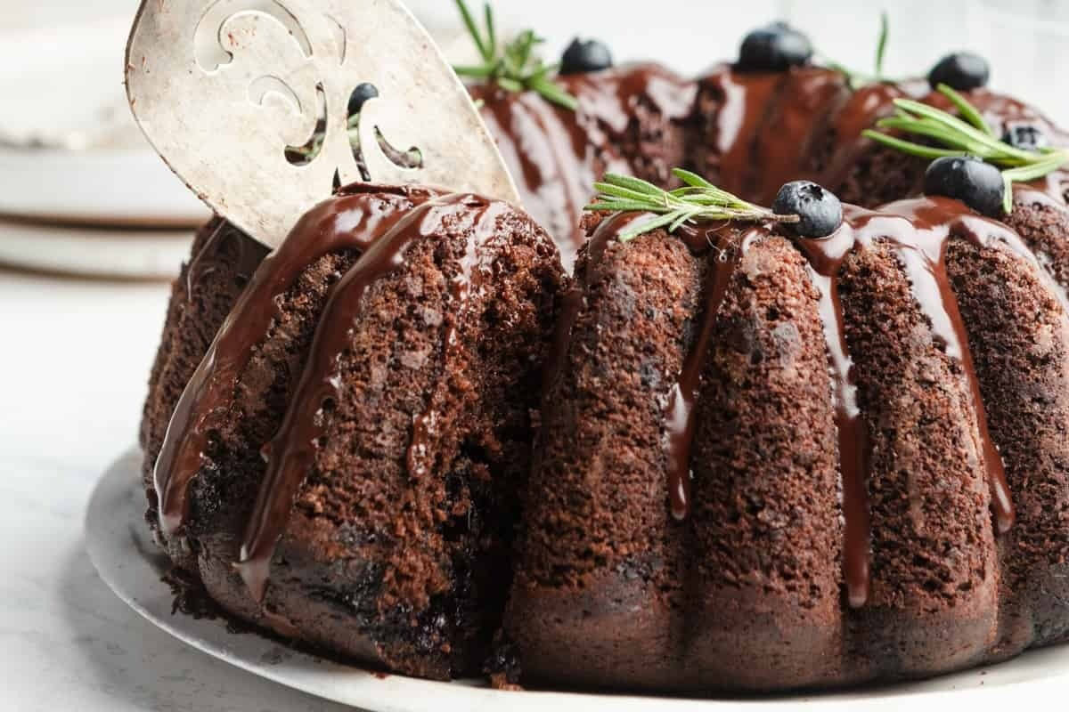 A chocolate bundt cake with chocolate glaze, garnished with blueberries and rosemary, has a slice being lifted out with a metal server.