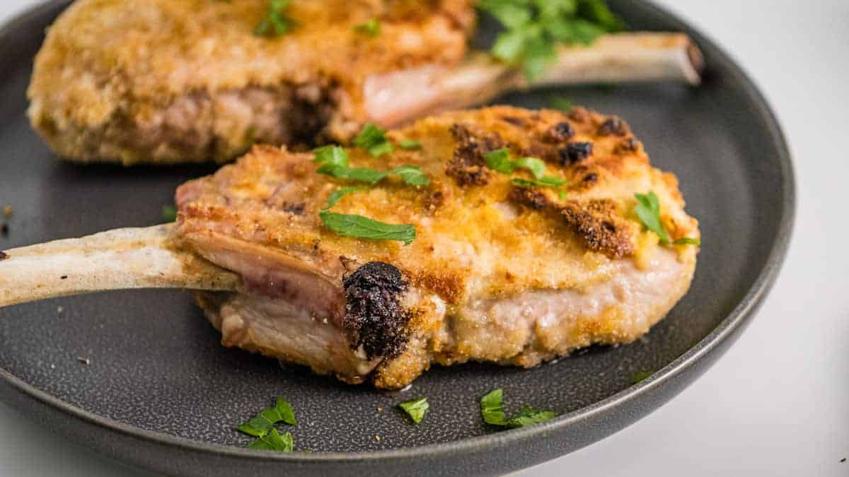 Two breaded bone-in pork chops garnished with chopped fresh herbs on a dark plate.
