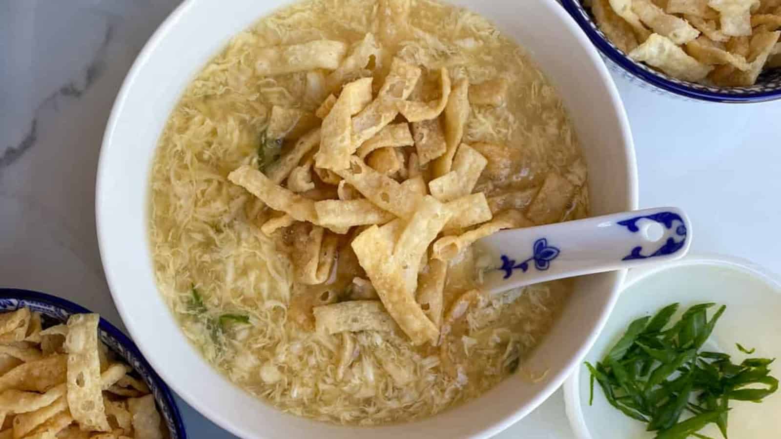 A bowl of egg drop soup topped with crispy fried wonton strips, with a spoon resting inside and side dishes of more wontons and chopped green onions nearby.