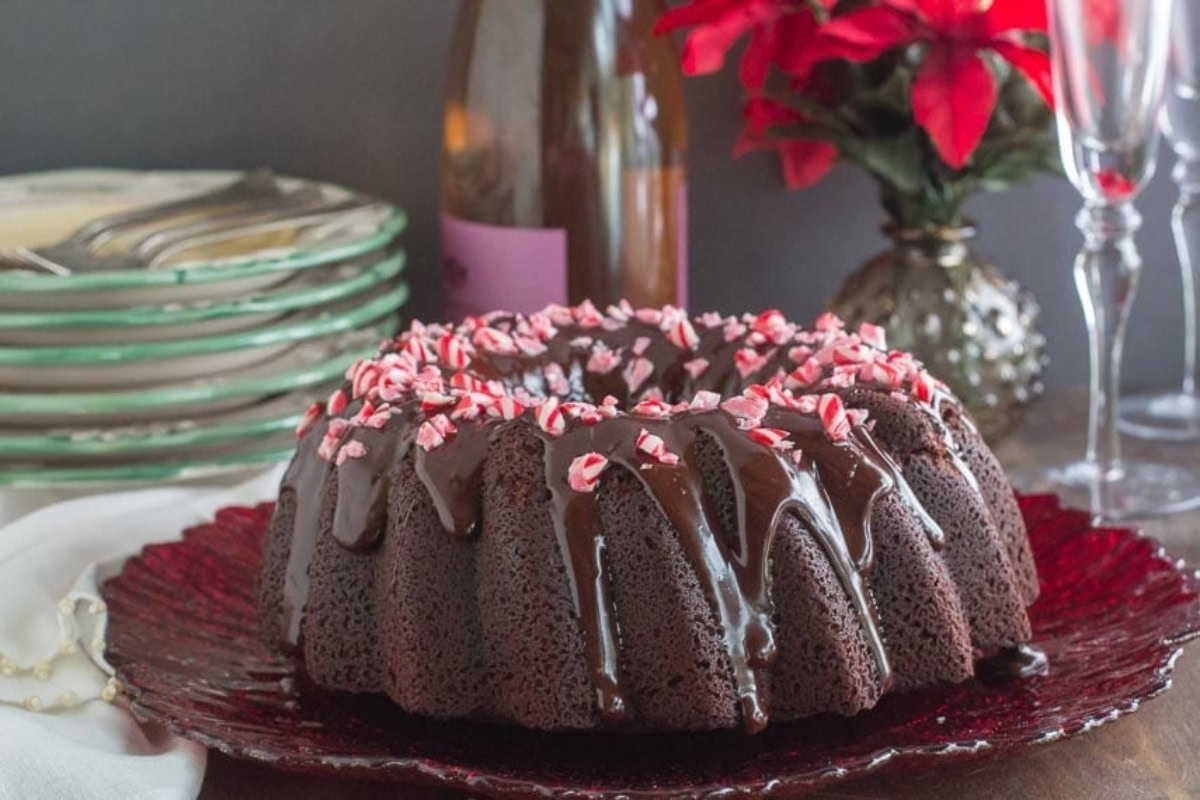 A chocolate bundt cake topped with chocolate glaze and crushed peppermint sits on a red plate, with stacked plates, a flower, and glasses in the background.