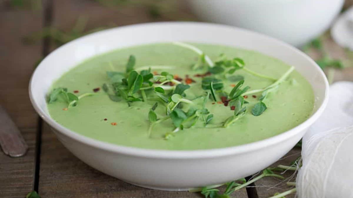 A white bowl filled with creamy green soup, garnished with fresh herbs and a sprinkle of red pepper flakes, placed on a wooden surface.