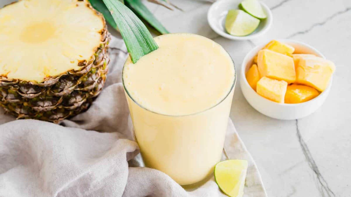 A glass of yellow smoothie garnished with a pineapple leaf, next to a halved pineapple, lime wedges, and a bowl of mango chunks on a white surface.