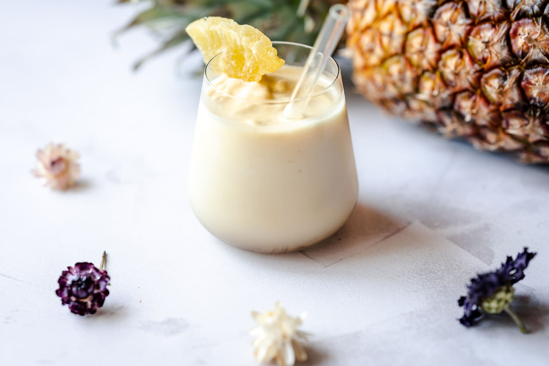A glass of creamy pineapple smoothie with a straw and pineapple garnish, placed on a white surface next to a whole pineapple and scattered dried flowers.