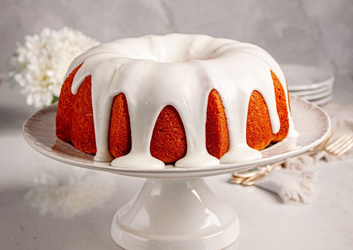 A bundt cake with white icing is displayed on a cake stand, with plates, forks, and a white flower in the background.
