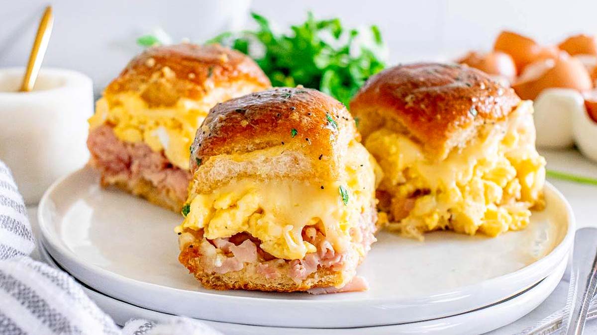 Three breakfast sliders with scrambled eggs, ham, and melted cheese served on a white plate, with greens and eggs in the background.