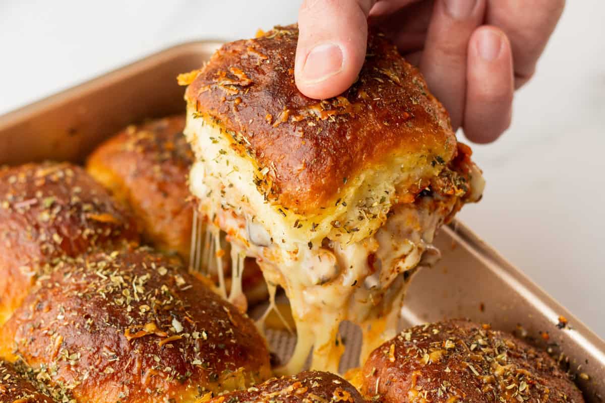 A hand lifts a cheesy, golden-brown baked slider from a tray of pull-apart sandwiches topped with herbs.
