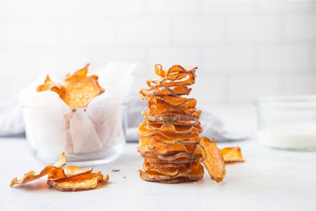 A stack of homemade sweet potato chips sits on a white surface, with a bowl of more chips and a glass jar in the background.
