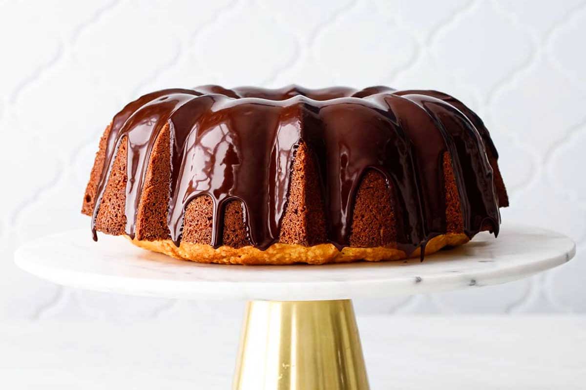 A chocolate-glazed bundt cake sits on a marble cake stand with a gold base against a white textured background.