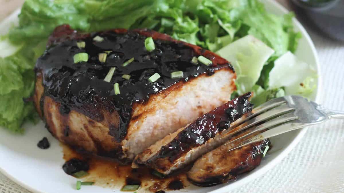 A thick pork chop topped with a dark glaze, garnished with chopped green onions, served with leafy green lettuce on a white plate.