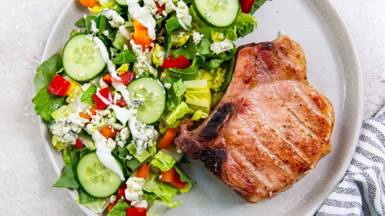 A grilled pork chop served on a plate with a mixed green salad topped with sliced cucumbers, chopped peppers, blue cheese, and ranch dressing.