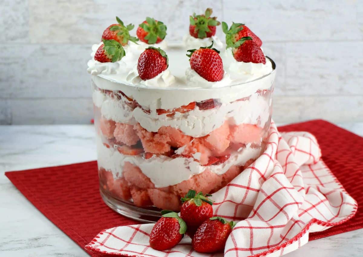 A trifle dessert with layers of whipped cream, pink cake, and strawberries in a glass bowl, topped with whole strawberries, sits on a red cloth with extra strawberries nearby.