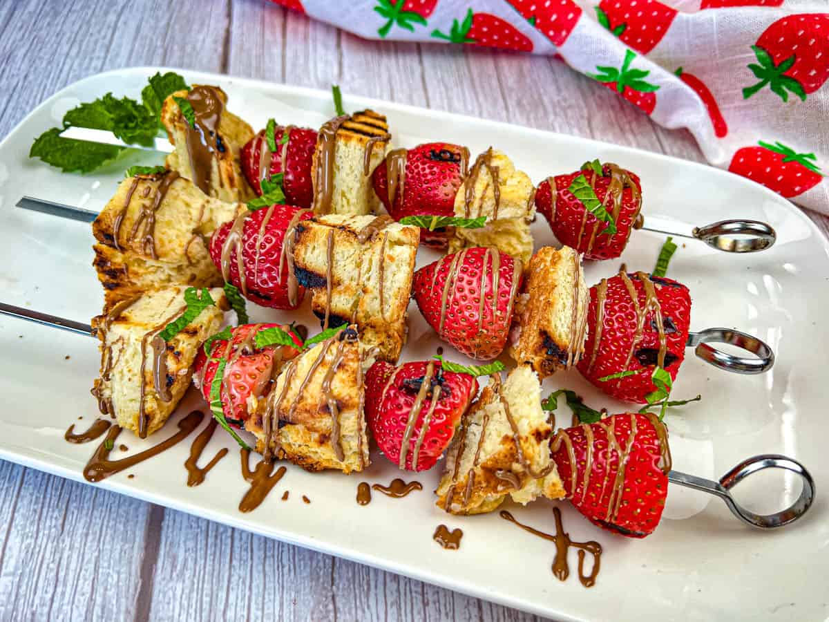 Skewers with alternating pieces of toasted bread and strawberries, drizzled with chocolate and garnished with mint, arranged on a white plate.