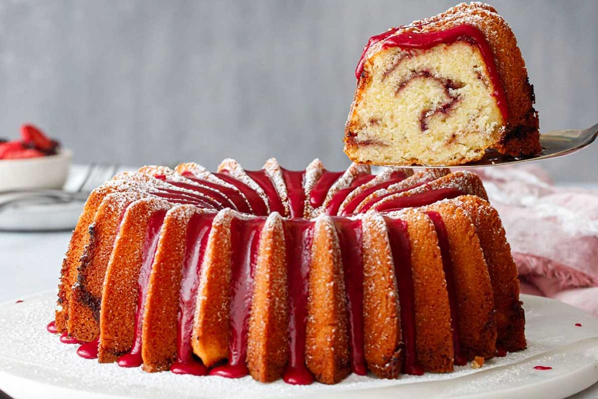 A sliced bundt cake with red glaze and powdered sugar on top, with one piece lifted to reveal a swirled filling inside.