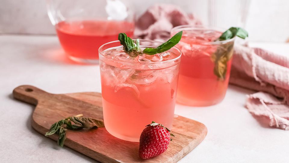 Two glasses of pink iced drink garnished with basil leaves, a fresh strawberry, and basil on a wooden board, with a pitcher and cloth in the background.
