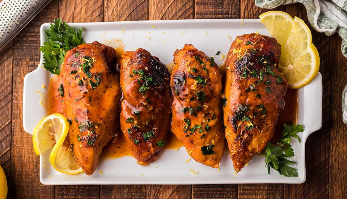 Four glazed chicken breasts garnished with chopped herbs are arranged on a rectangular white plate with lemon slices and parsley on a wooden surface.