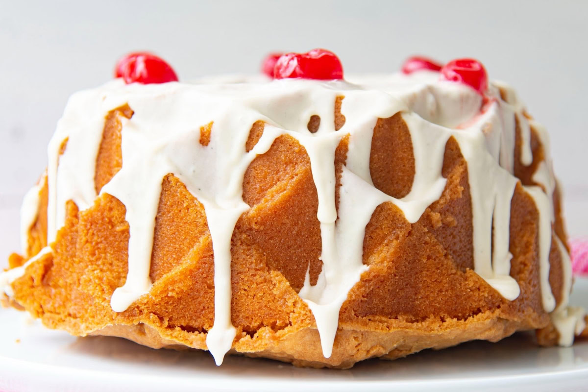 A bundt cake with white icing drizzled on top, decorated with red cherries, sits on a white plate.