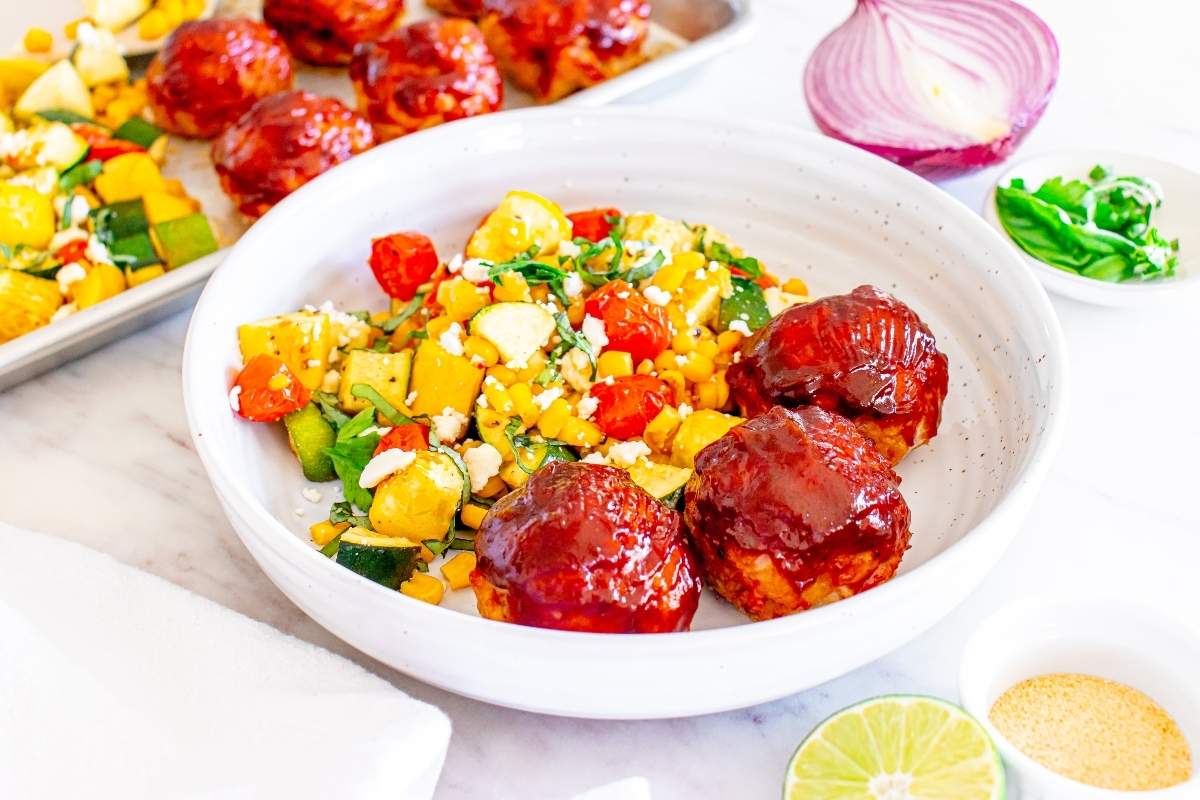 A white bowl with three glazed meatballs and a colorful vegetable salad, surrounded by ingredients like onion, herbs, a lime half, and a baking sheet of more meatballs.