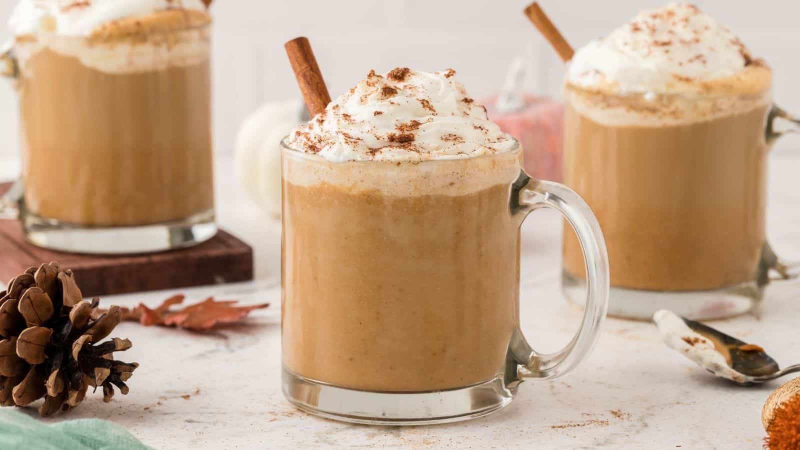 Three glass mugs of pumpkin spice latte topped with whipped cream and cinnamon sticks sit on a white surface, surrounded by autumn-themed decor.