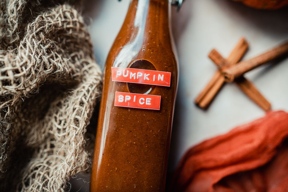 A glass bottle labeled Pumpkin Spice with a brown liquid inside, next to a piece of burlap fabric and cinnamon sticks.