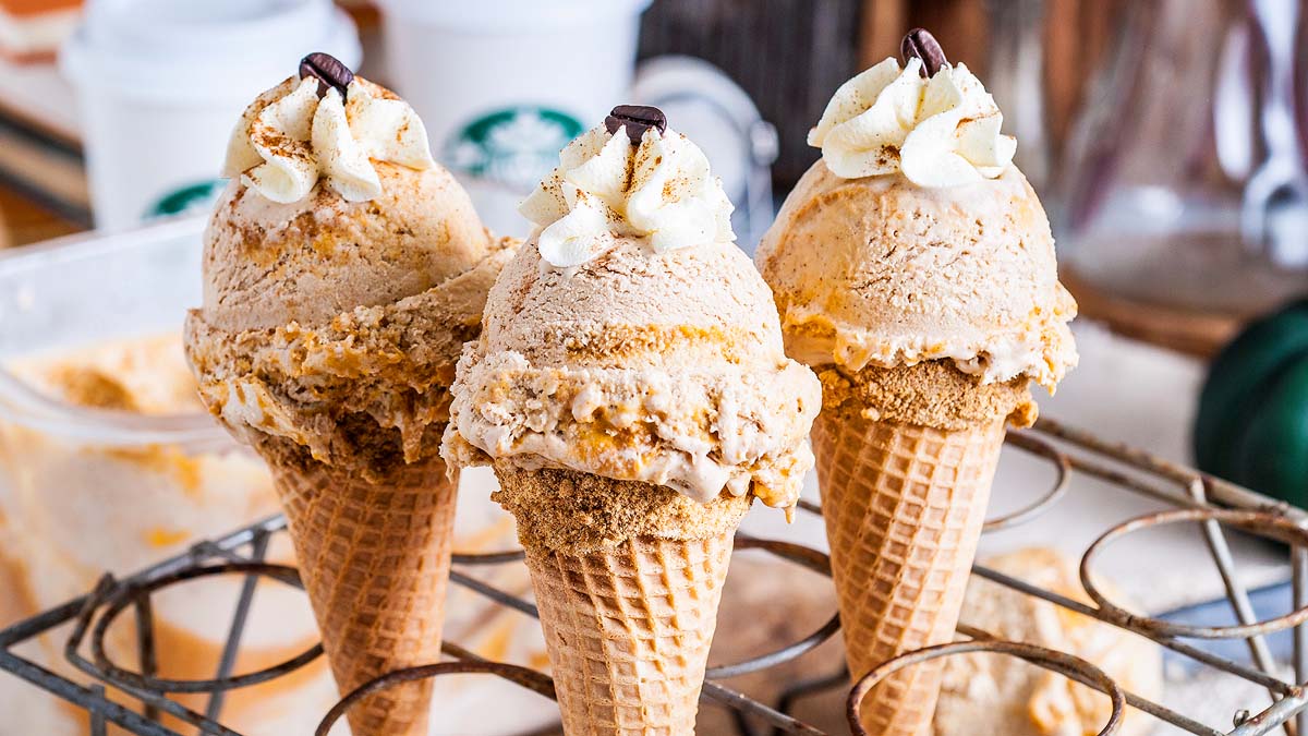 Three ice cream cones with scoops of light brown ice cream, topped with whipped cream and a coffee bean, are displayed on a metal rack. Starbucks cups are blurred in the background.