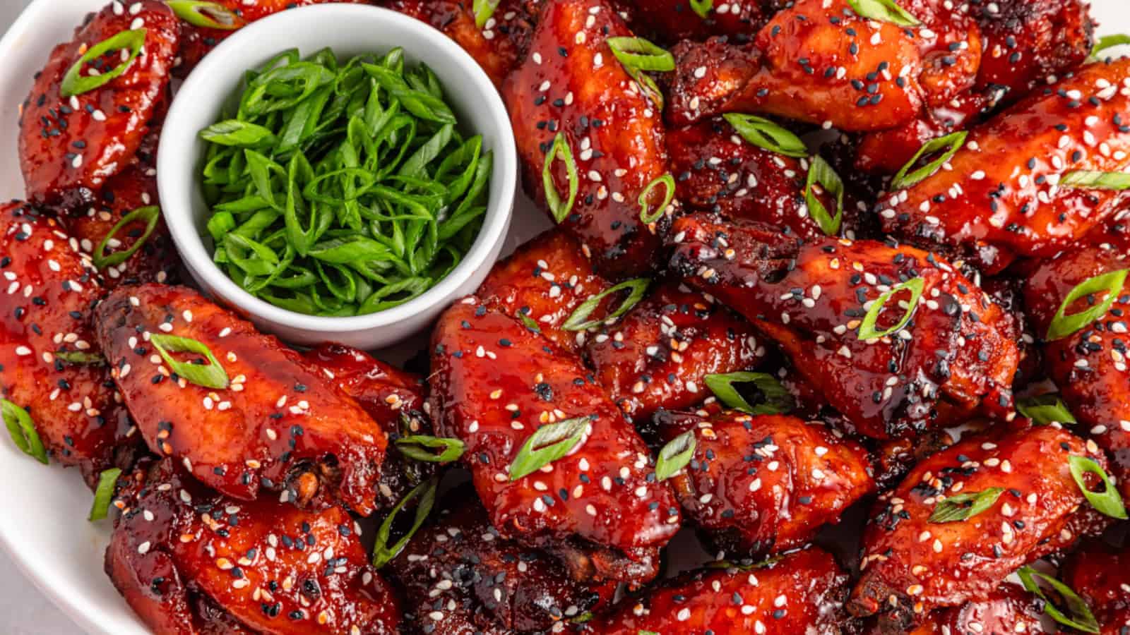 A plate of glazed chicken wings garnished with sesame seeds and sliced green onions, served with a small bowl of chopped green onions.