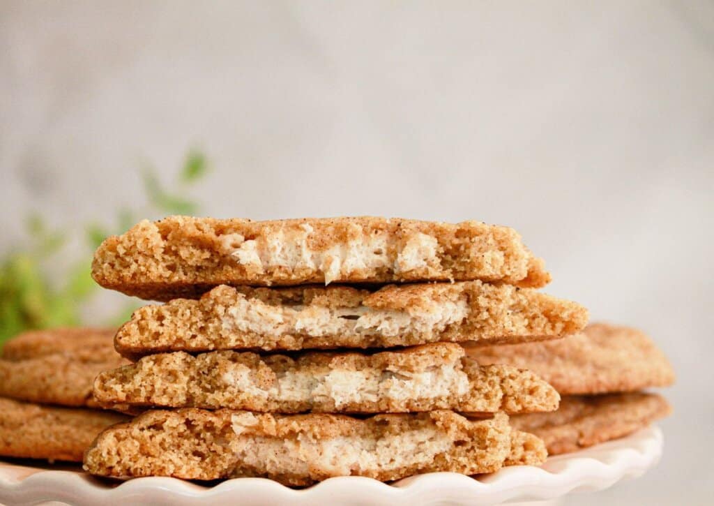 A stack of cinnamon cookies broken in half, showing a creamy white filling, displayed on a white scalloped plate.