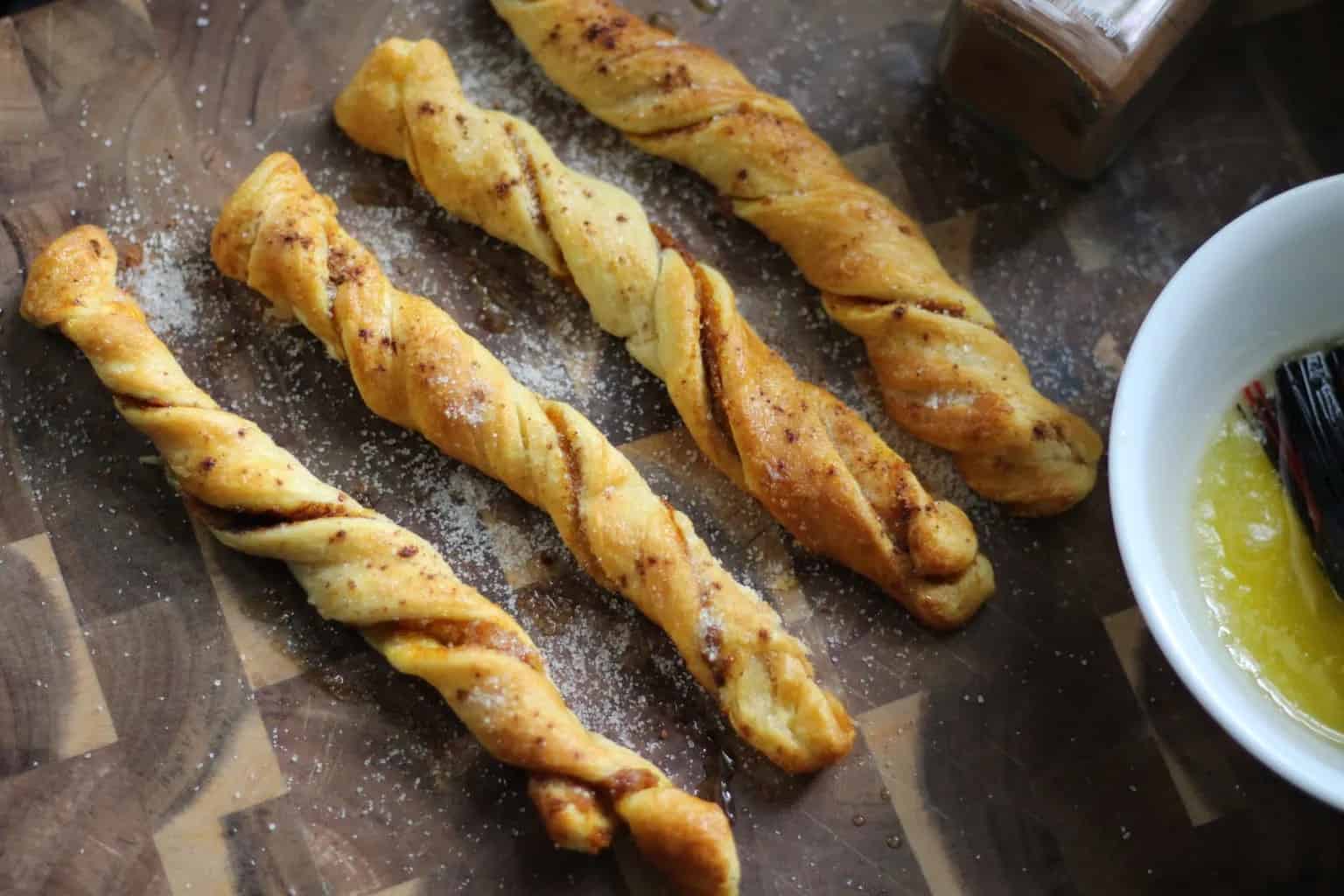 Four twisted pastry sticks dusted with sugar are arranged on a wooden surface next to a bowl containing melted butter and a brush.
