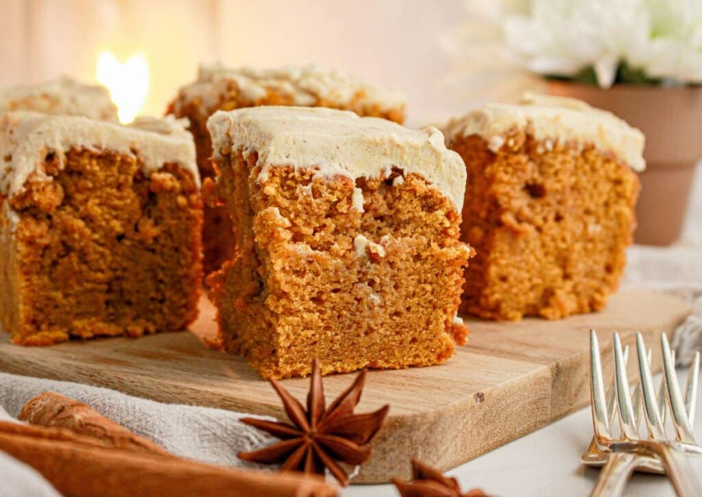 Four pieces of frosted pumpkin cake are arranged on a wooden board, with star anise, cinnamon sticks, forks, and a potted flower visible nearby.