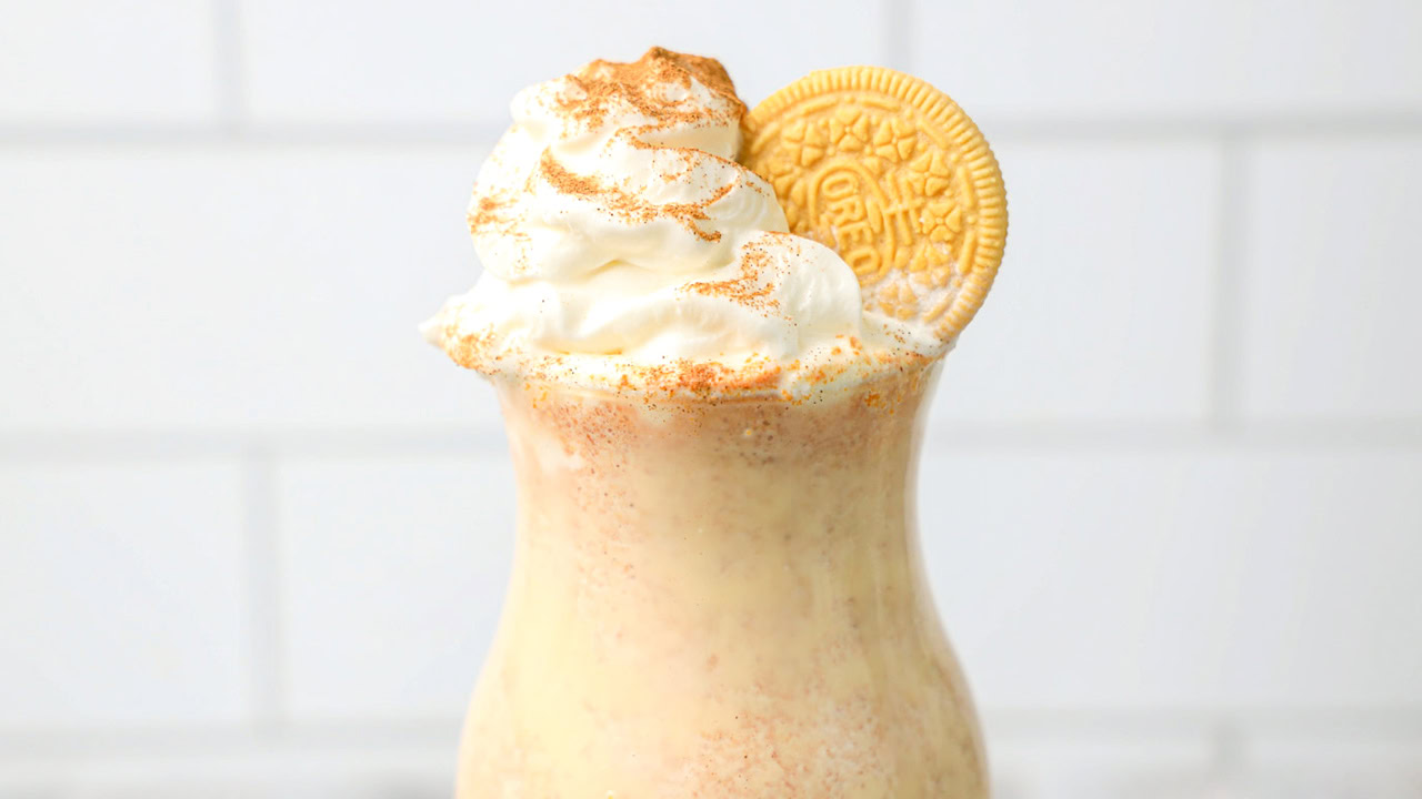 A milkshake topped with whipped cream, cinnamon, and a golden sandwich cookie, served in a glass against a white tiled background.
