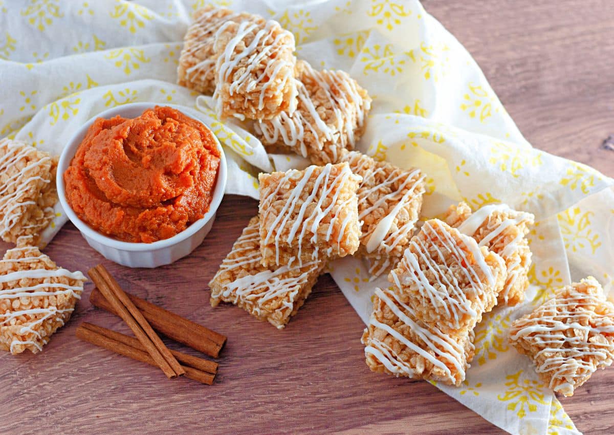 Rectangular rice cereal treats drizzled with white icing are arranged next to a bowl of orange dip and cinnamon sticks on a wooden surface with a patterned cloth.