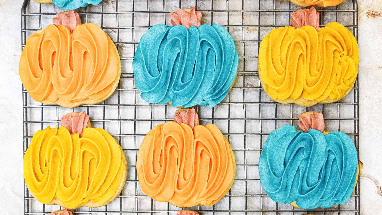 Pumpkin-shaped cookies with orange, blue, and yellow frosting are arranged on a cooling rack.