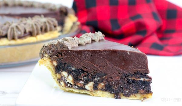 A slice of chocolate pie with a thick chocolate filling, nut pieces, and a decorative chocolate topping, served on a white plate.