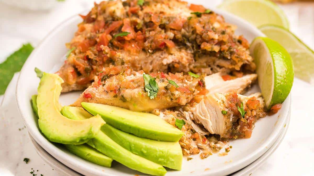 A plate of baked chicken topped with tomato salsa, served with sliced avocado and a lime wedge.