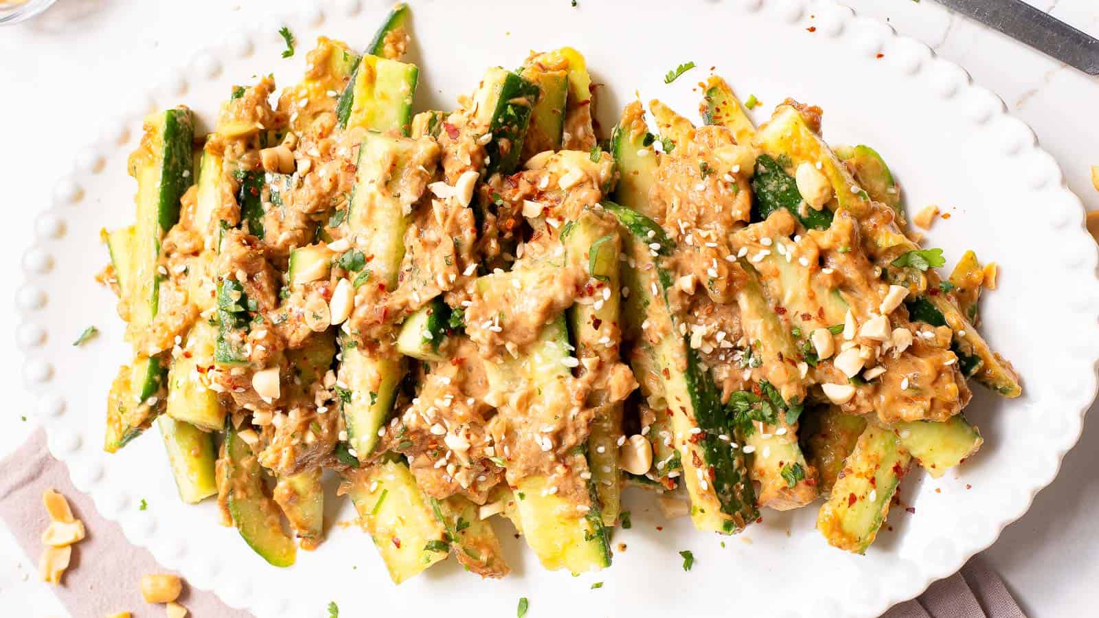 Sliced cucumbers arranged on a white plate, topped with a creamy peanut sauce, chopped herbs, and crushed peanuts.