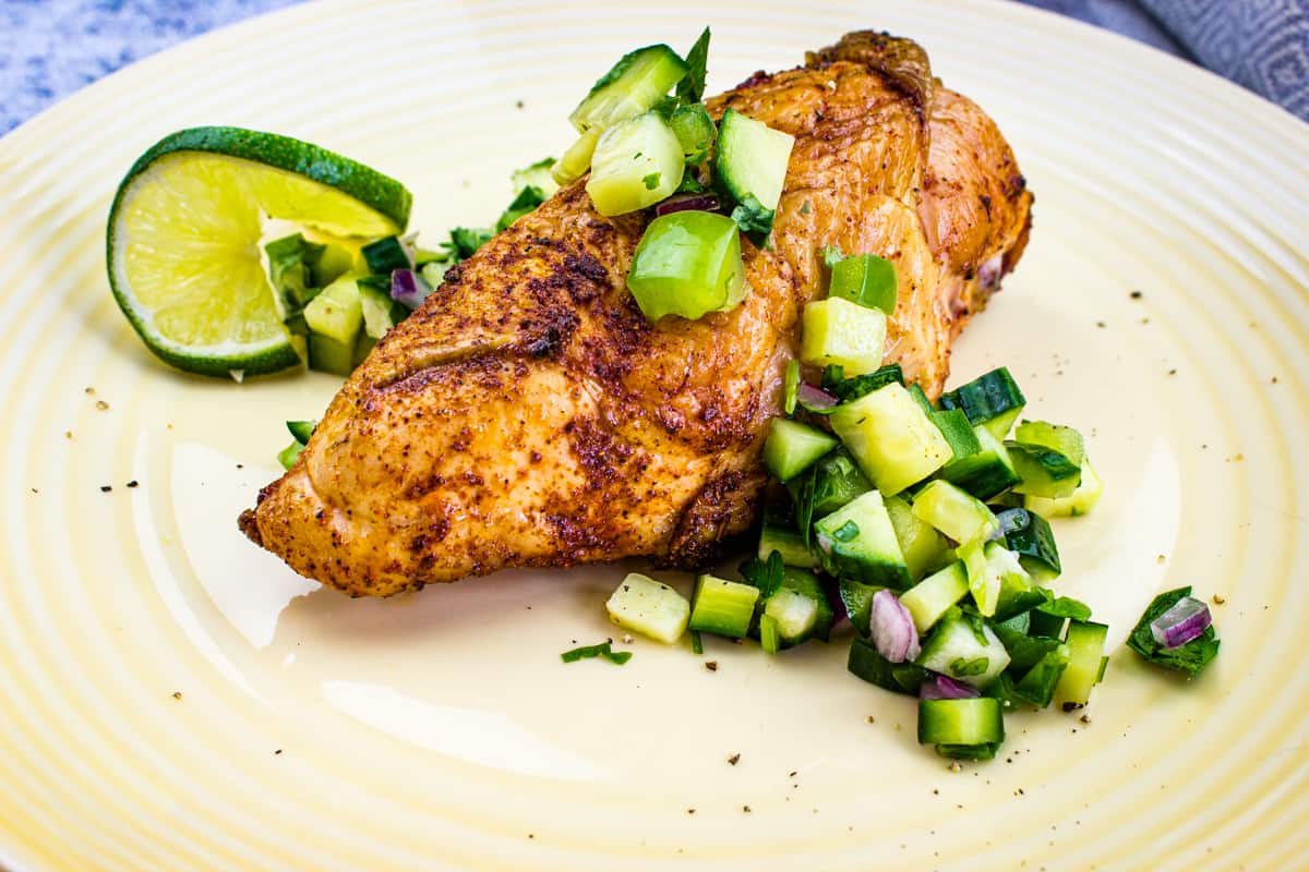 Seasoned grilled chicken breast topped with chopped cucumber and red onion salsa, garnished with a lime wedge on a cream-colored plate.