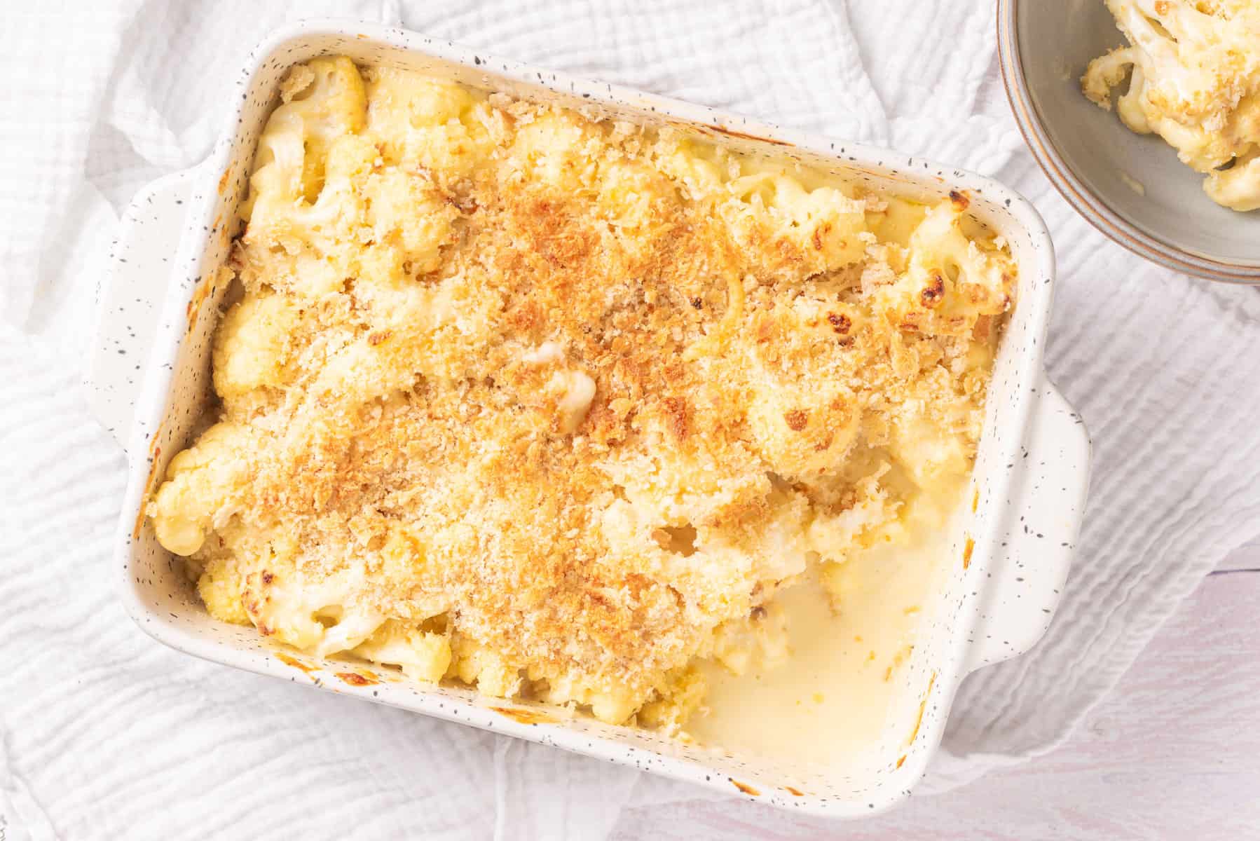 Baked cauliflower gratin with a golden breadcrumb topping in a white rectangular dish, with a portion served out on a nearby plate.