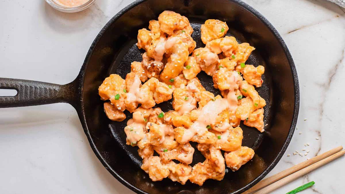 A black skillet filled with pieces of battered and sauced cauliflower, topped with a creamy dressing and garnished with chopped herbs.