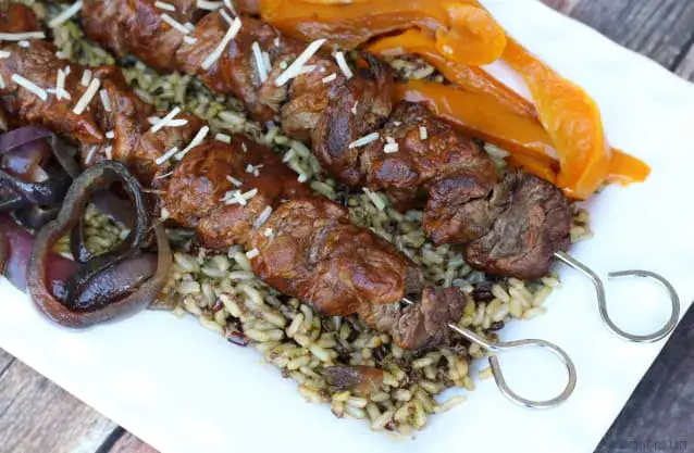 Skewers of grilled meat served on a bed of rice with sliced orange bell peppers and red onions on a white plate.