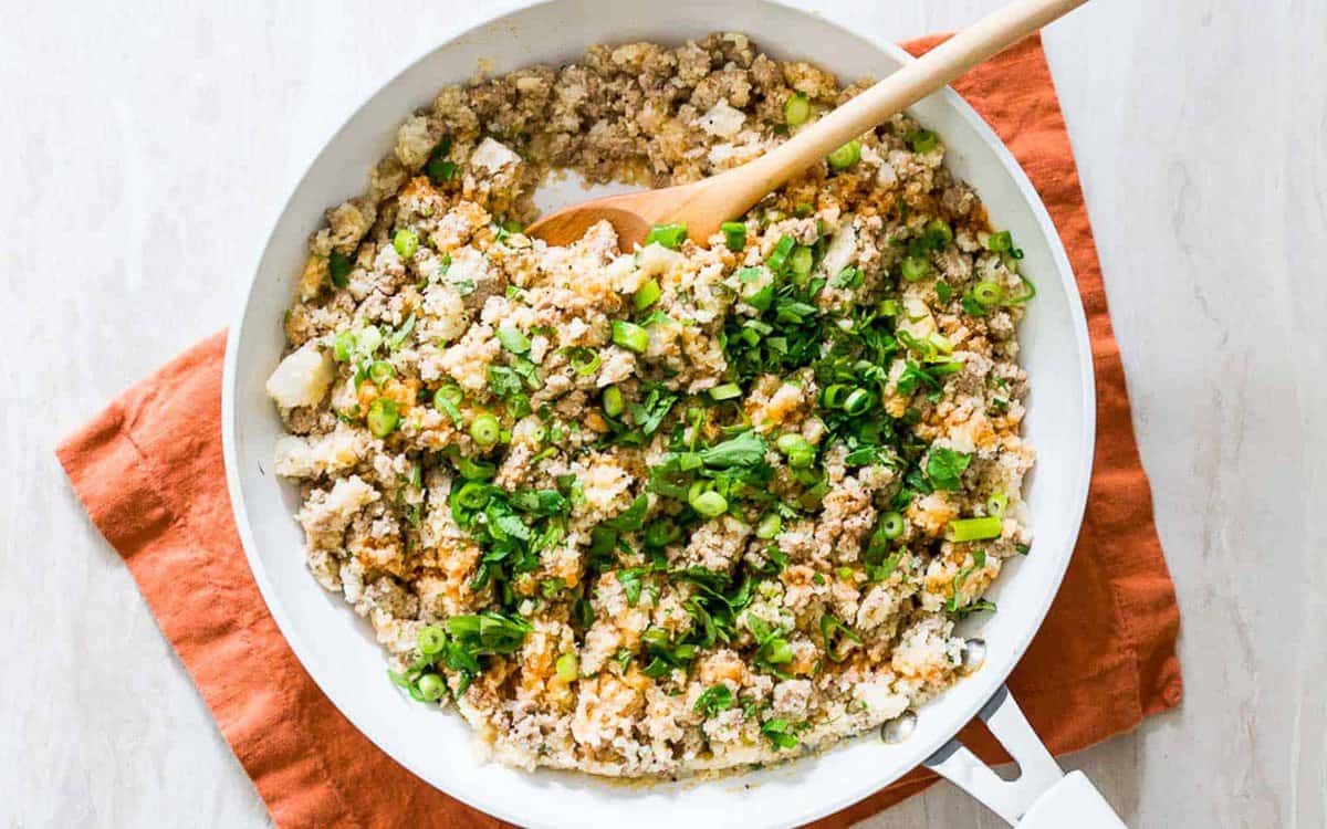 A skillet of ground meat, riced cauliflower, chopped green onions, and herbs, with a wooden spoon on an orange cloth napkin.