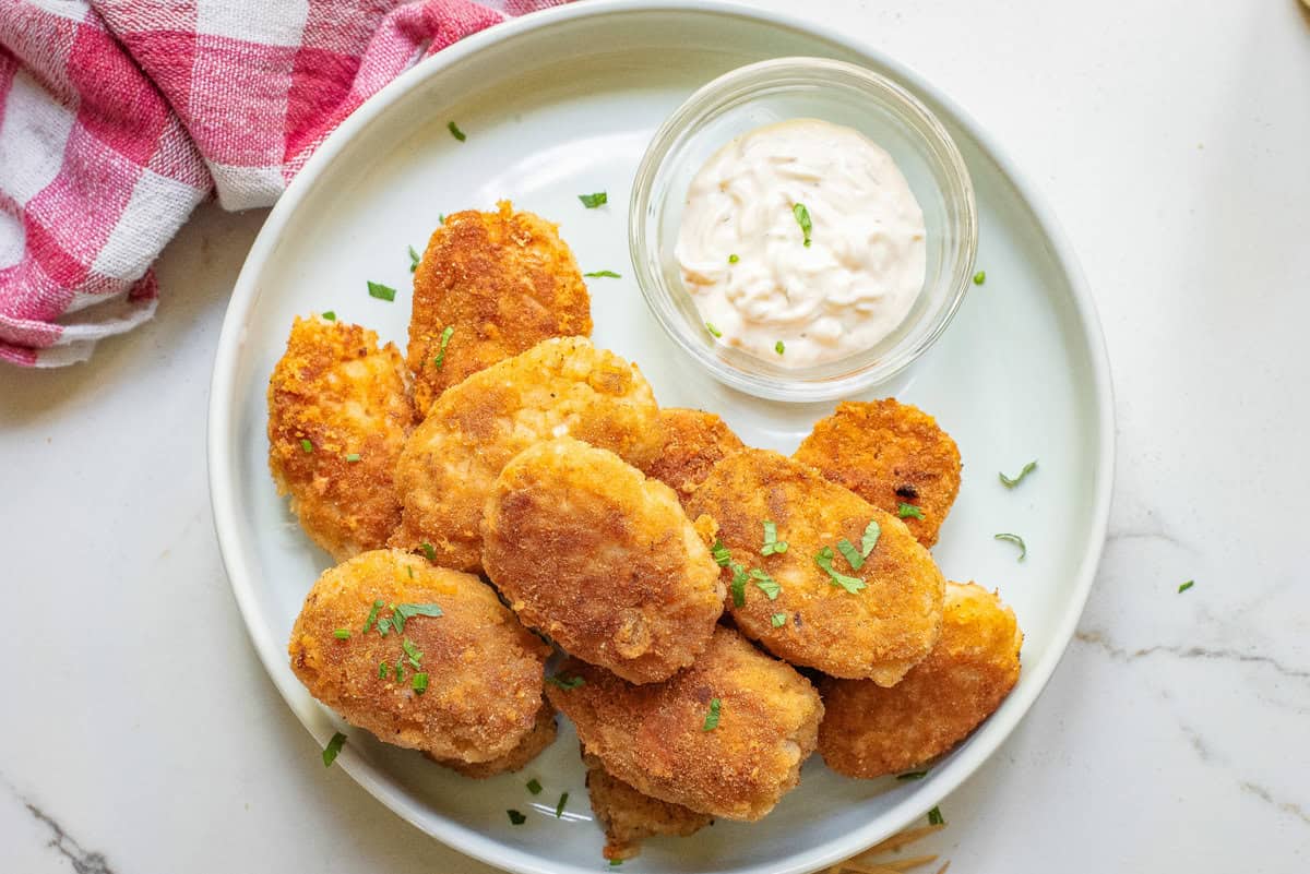 A plate of breaded chicken nuggets garnished with herbs, served with a small bowl of dipping sauce on a white surface. A red and white cloth is partially visible in the corner.