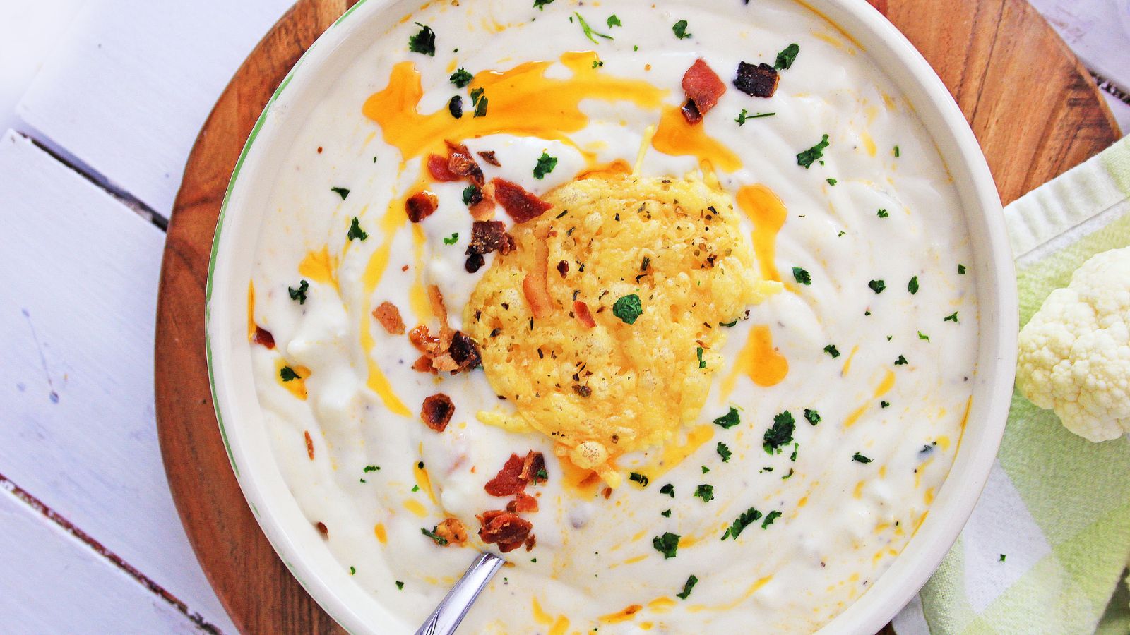 A bowl of creamy soup garnished with melted cheese, bacon bits, and chopped herbs, with a spoon resting inside. A piece of cauliflower is visible beside the bowl.