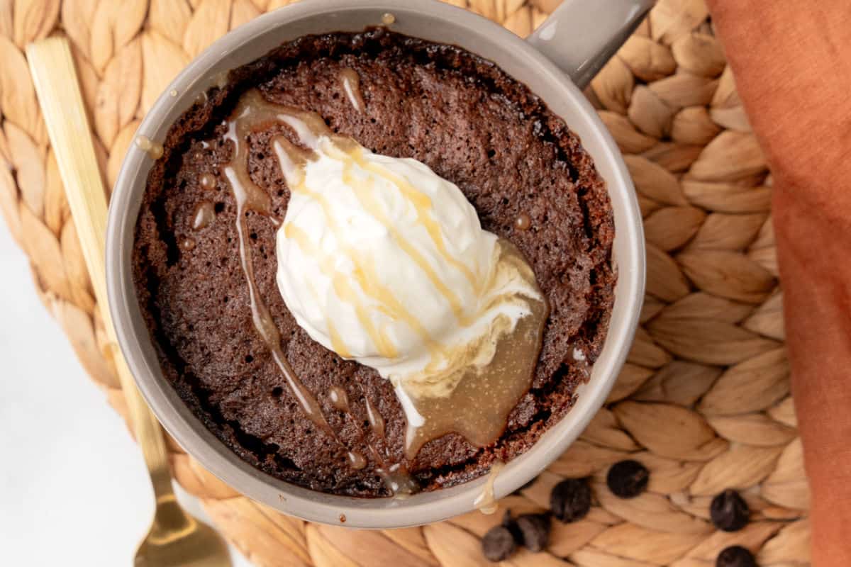 A chocolate mug cake topped with a scoop of vanilla ice cream and caramel sauce, placed on a woven mat with a gold spoon nearby.