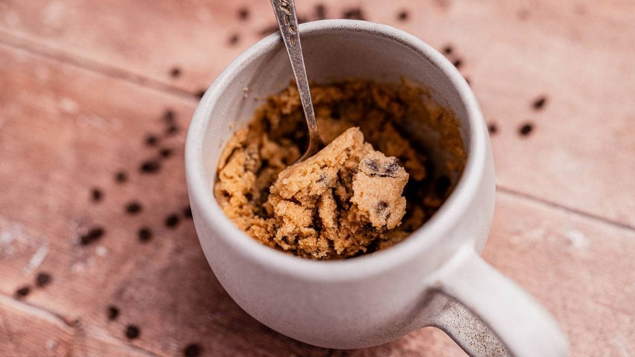 A white mug with a spoon inside, filled with chocolate chip cookie dough, sits on a wooden surface with scattered chocolate chips around it.