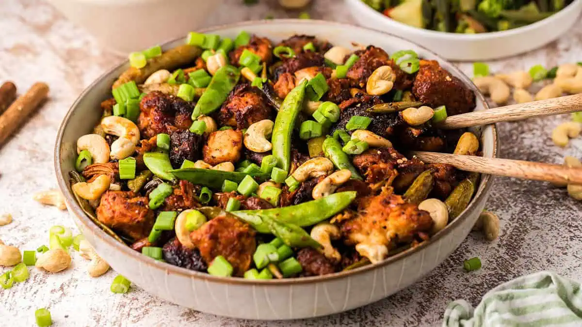 A bowl of stir-fried chicken with snap peas, scallions, cashews, and sauce, with chopsticks resting on the edge.
