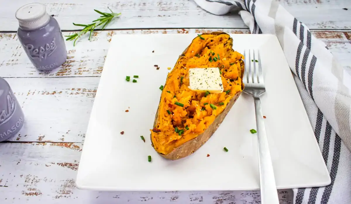 A baked sweet potato half topped with butter and chives sits on a white plate with a fork, next to salt and pepper shakers and a striped napkin.
