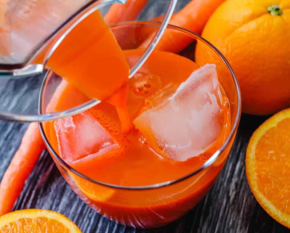 A glass of orange juice with ice cubes is being poured from a jug, surrounded by fresh carrots and orange slices on a wooden surface.