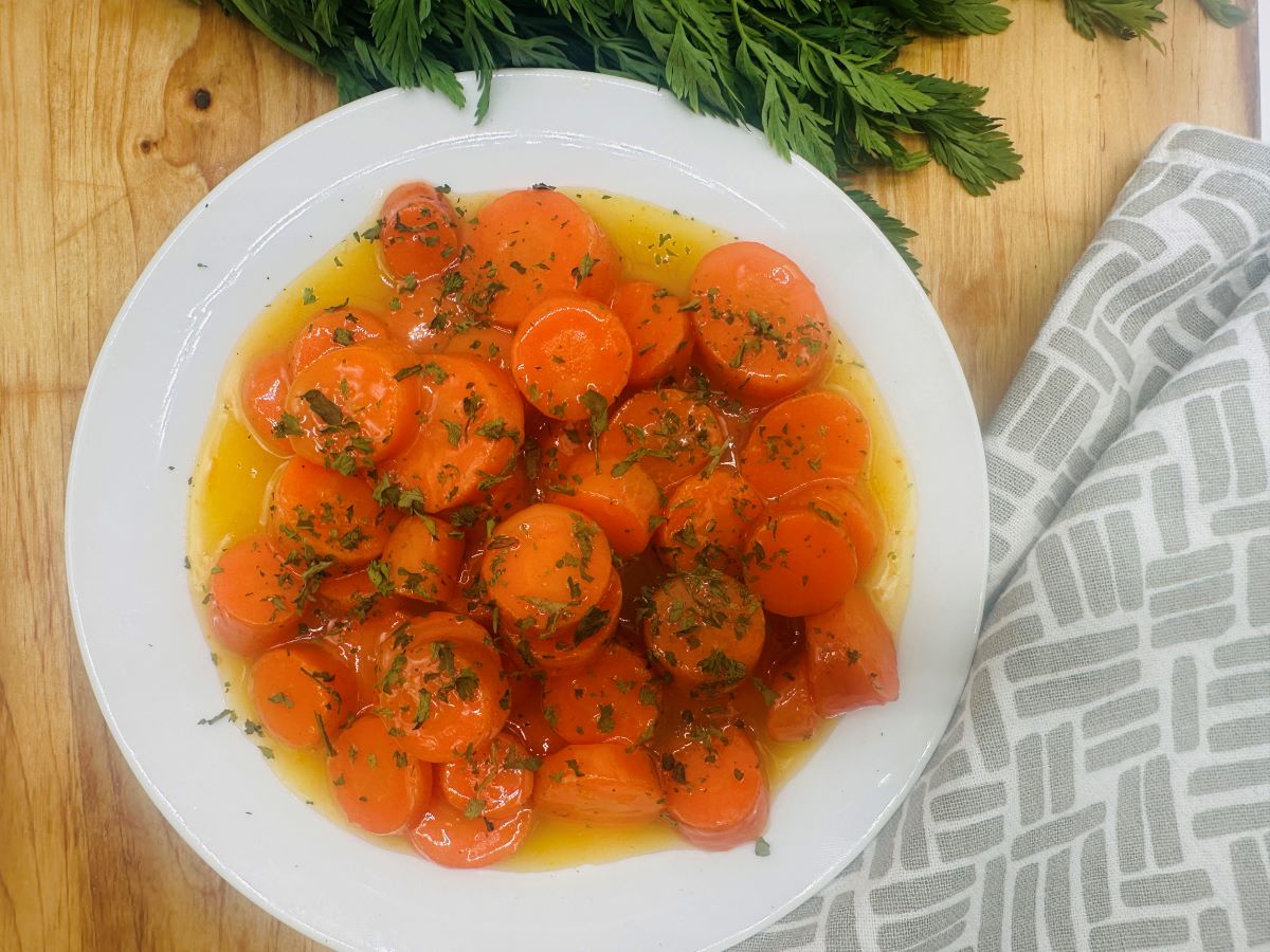 A white plate of sliced cooked carrots in a yellow glaze, garnished with chopped herbs, sits on a wooden surface near a gray patterned cloth and carrot greens.
